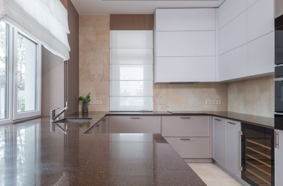 Minimalist interior kitchen design with stone countertop of marble color with sink and chrome plated faucet lying on cupboard with built in stove and electric appliances in daylight